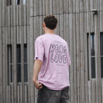 Person wearing a pink t-shirt with 'WEAR LOVE' text, standing in front of a wooden building.