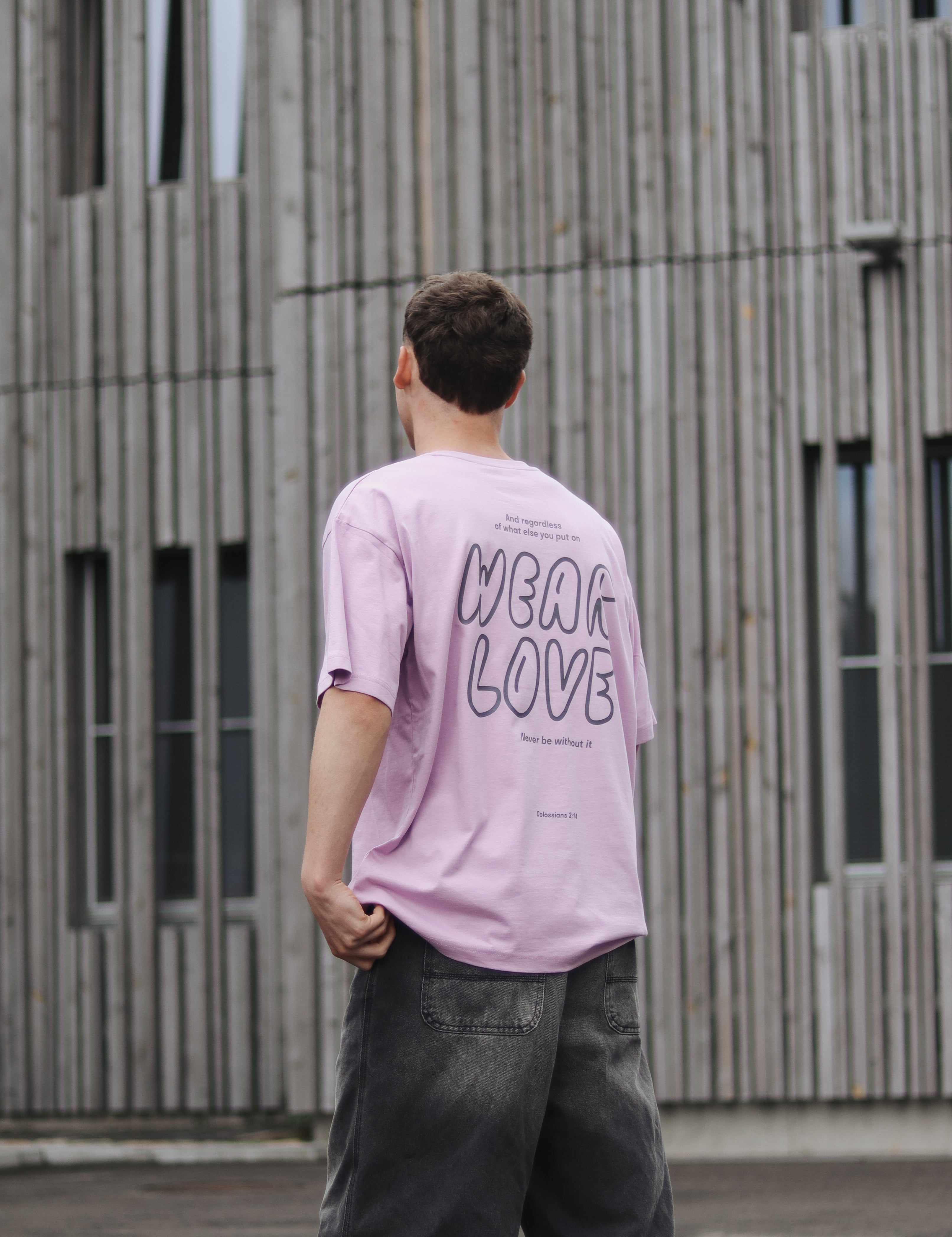 Person wearing a pink t-shirt with 'WEAR LOVE' text, standing in front of a wooden building.