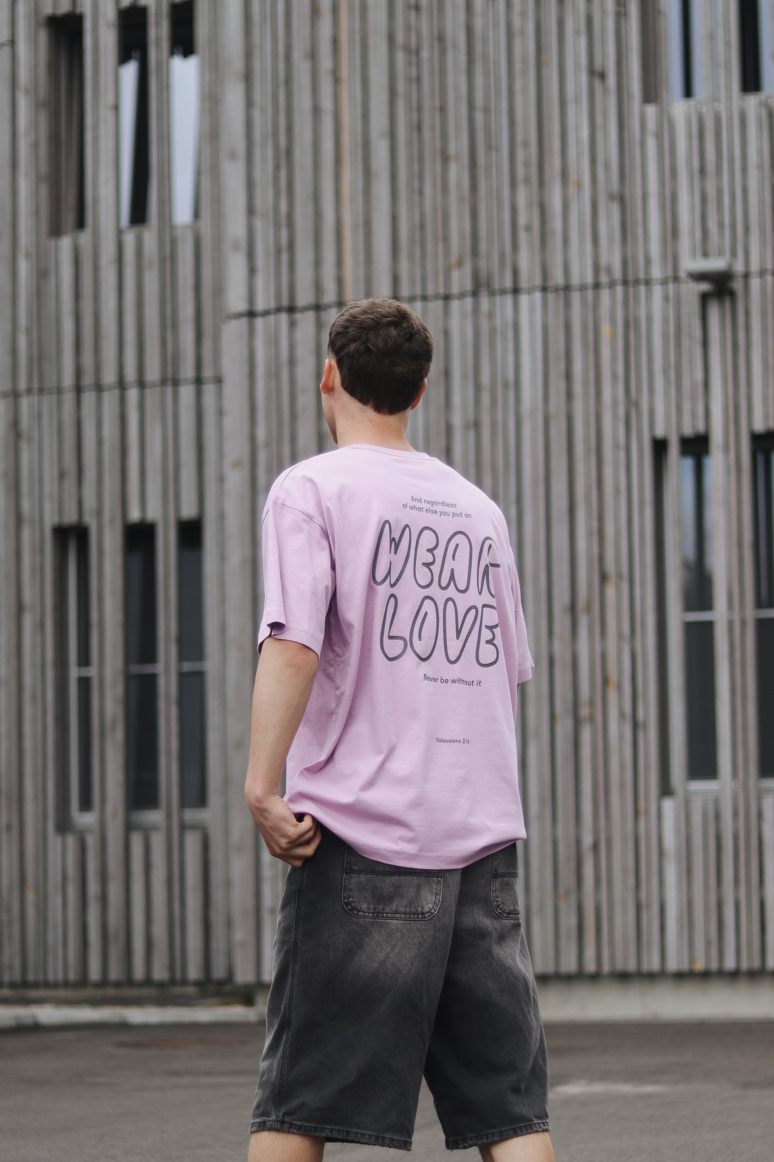 Person wearing a pink t-shirt with 'WEAR LOVE' text, standing in front of a wooden building.
