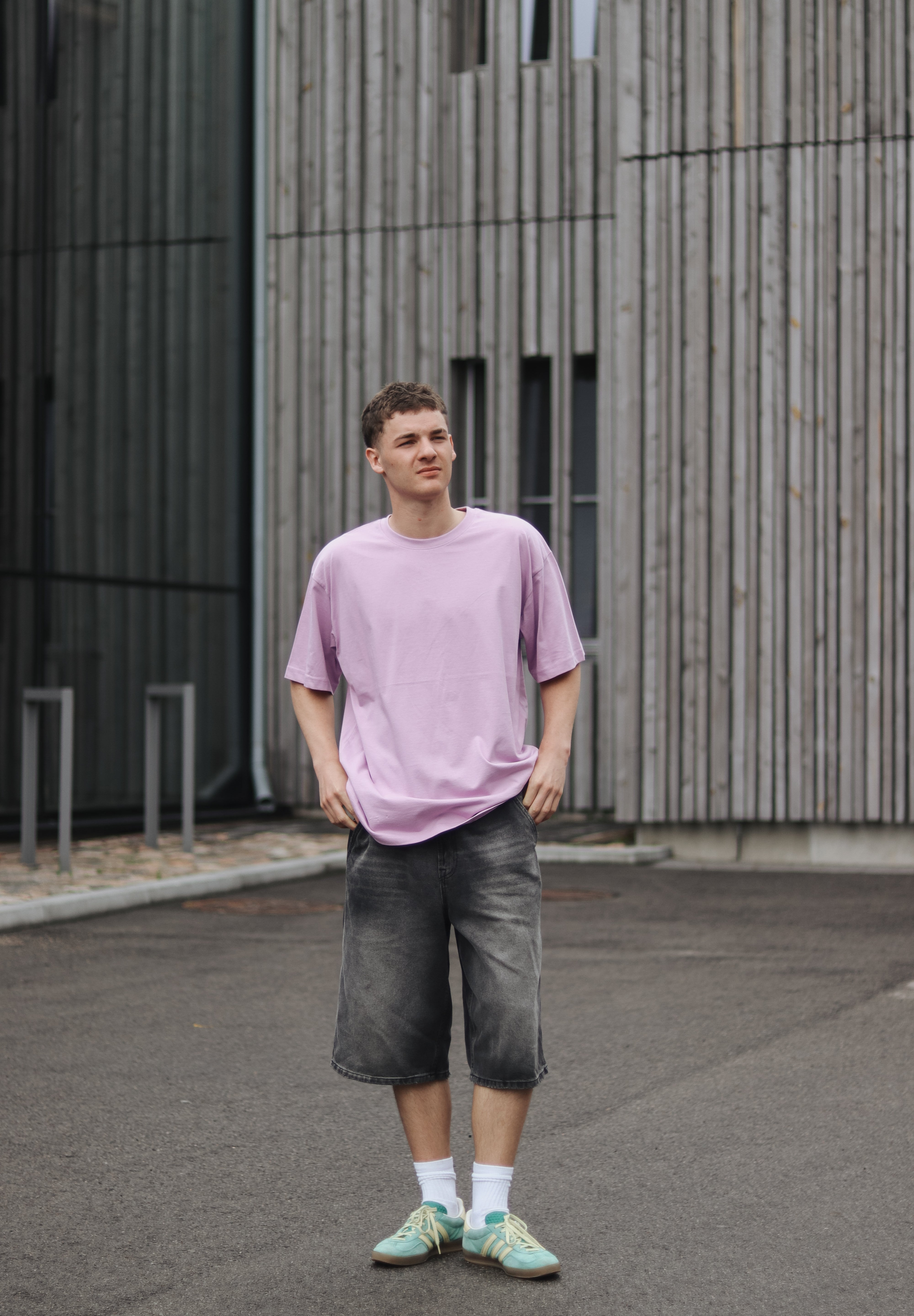 Person wearing a light purple t-shirt and dark shorts standing in front of a modern building.